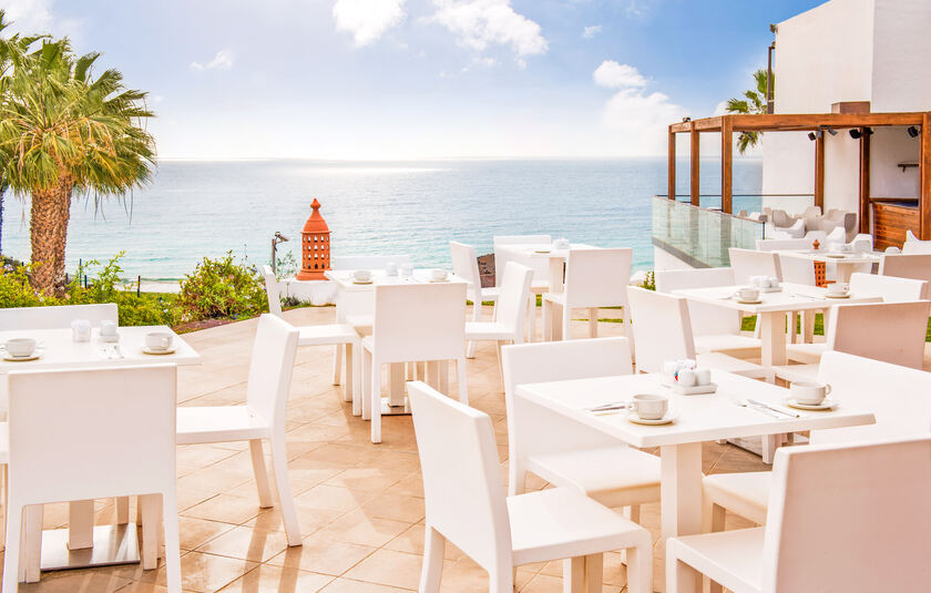 Udendørs restaurantterrasse på hotellet Esencia de Fuerteventura by Princess, præget af de lyse hvide møbler og den direkte havudsigt.